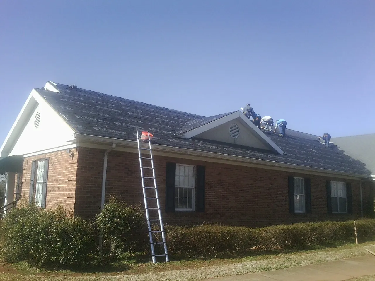 Crew tearing shingles off a brick ranch roof with a ladder set — Metal Roof Repair work by our crew in Wartburg
