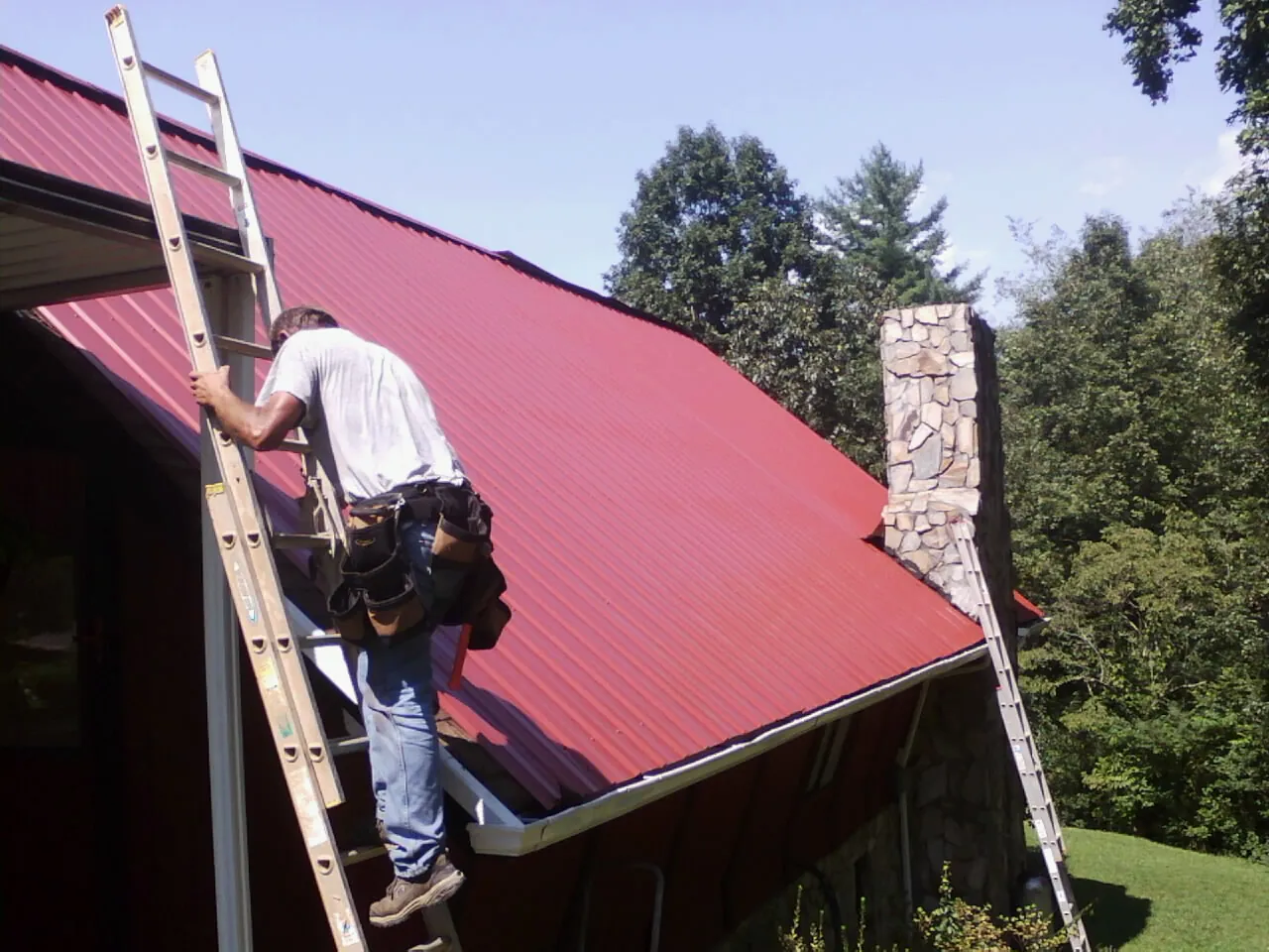 Roofer on a ladder beside a red metal roof and stone chimney — Soffit & Fascia Repair by Roofing Brothers in Wartburg